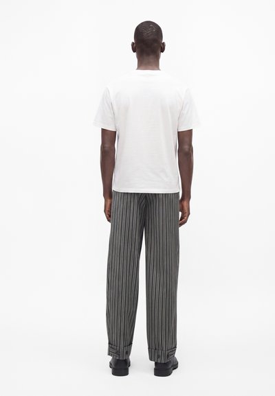 Homme debout de dos portant une chemise blanche unie à manches courtes, un pantalon large à rayures verticales noires et blanches, et des chaussures noires.