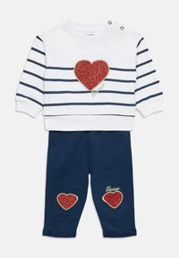 Άσπρο φούτερ με ναυτικό ρίγες, κόκκινο πατς καρδιά με γκλίτερ και λογότυπο "Guess". Σκούρο μπλε παντελόνι με δύο ταιριαστά πατς καρδιά και κέντημα.