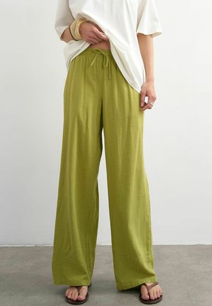 Personne portant un pantalon ample vert olive avec cordon, un haut blanc oversize et des tongs marron, debout devant un mur clair uni.