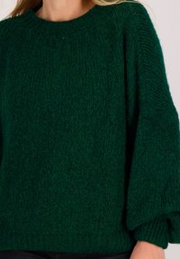 Vrouw draagt een donkergroene, grove gebreide trui met lange, licht opgepofte mouwen en een ronde hals.