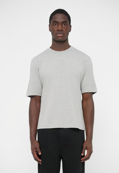 T-shirt gris à manches courtes avec un col rond et un subtil accent circulaire. Le tissu semble doux et texturé. Porté avec un pantalon noir.