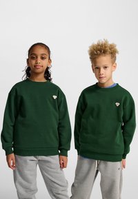 Gröna sweatshirts med rund halsringning och ribbade muddar, matchas med ljusgråa sweatpants; modeller visar en liten logotyp på sweatshirts.