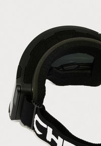 Gafas de esquí negras con forro de espuma, marco flexible y una correa blanca con logotipo. Las lentes tienen un ligero tinte y el diseño incluye un panel lateral.