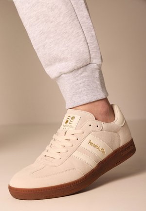 Sneaker beige in suede con accenti crema e design retro. Presenta lacci piatti, suola in gomma testurizzata e branding dorato sulla linguetta.