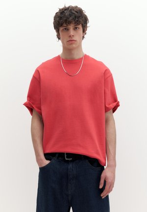 Ung mand med krøllet hår iført en løs rød t-shirt med opkørte ærmer, mørke bukser, sølvkæde om halsen, hænderne i lommerne, neutral baggrund.