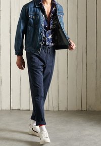 Chaqueta de denim, camisa de estampado, pantalones marinos, zapatillas blancas con detalles rojos. El atuendo presenta un aspecto casual con estilos y colores en capas.