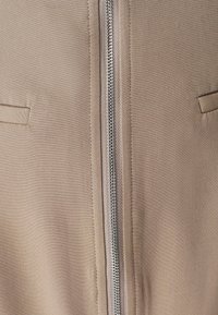 Chaqueta marrón claro con cremallera metálica frontal, dos bolsillos verticales y textura suave. Diseño minimalista con líneas limpias.