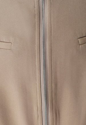 Jasnobrązowa kurtka z metalowym zamkiem z przodu, dwoma pionowymi kieszeniami i gładką fakturą. Minimalistyczny design z czystymi liniami.