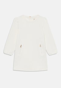 Hvit kjole med lange ballongsleeves, teksturert stoff og to frontlommer med glidelås. Rund hals med liten sløyfe til lukking bak.