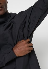 Czarna wodoodporna kurtka z wysokim kołnierzem, zamykana na zamek błyskawiczny kieszeń z przodu oraz regulowanym mankietem. Teksturowana tkanina o eleganckim, minimalistycznym designie.