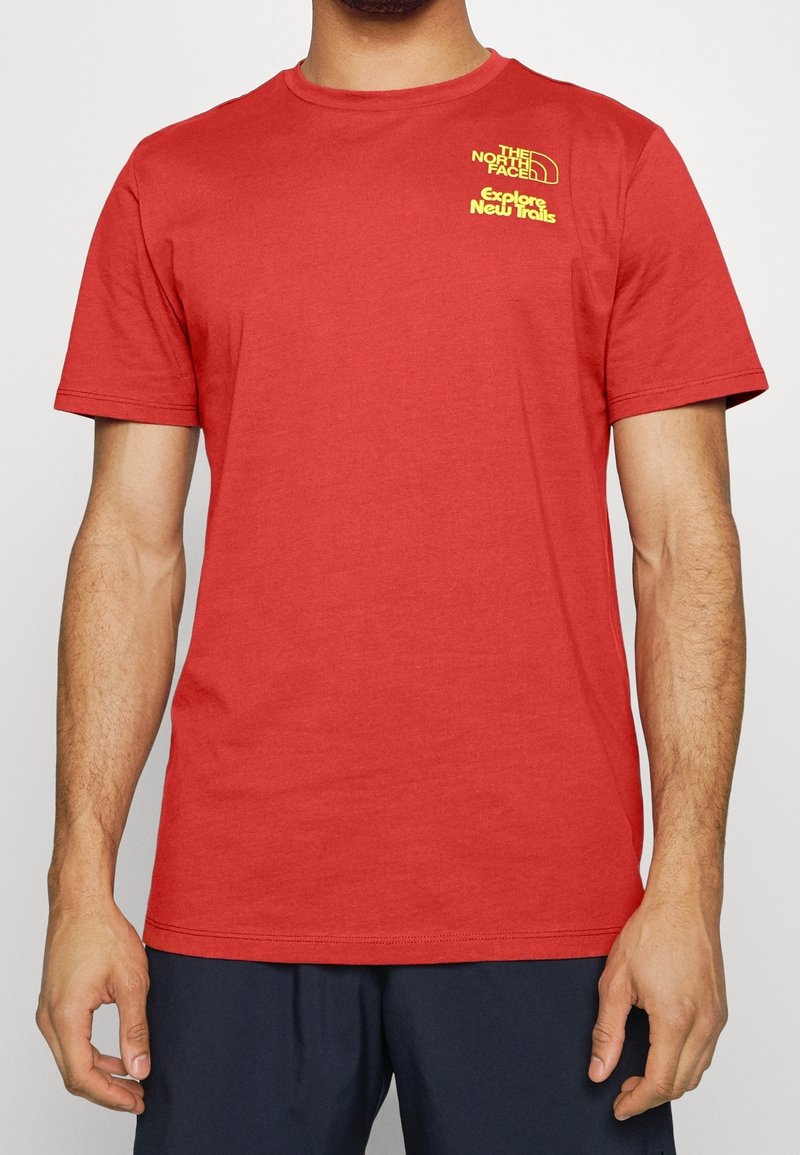 Camiseta de algodón roja con cuello redondo y mangas cortas. Presenta un logo y texto amarillos en la parte superior izquierda. Diseño sencillo, textura suave.