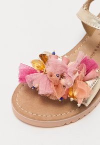 Sandale en simili cuir doré ornée d'une décoration florale en tulle rose et pêche avec des perles, reposant sur une semelle en suède beige avec des accents de couture.
