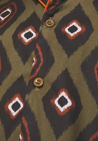 Gros plan sur un tissu vert olive avec de grands motifs losanges noirs, rouges et blancs et des boutons marron sur l'avant du vêtement.