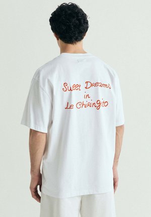 Hombre con cabello rizado y oscuro lleva una camiseta blanca holgada con texto en naranja que dice "Sweet Dreams in Le Chiringito" en la espalda.