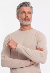 Beige pitkähihaine paita pyöreällä kauluksella, valmistettu pehmeästä kankaasta. Mallilla on harmaat hiukset ja parta, kädet ristissä.