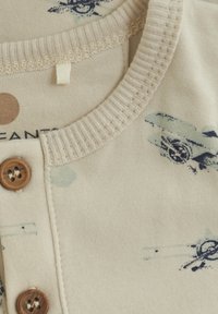 Beige bomullskjorta med en texturerad ribbad halsringning, med träknappar och ett blått abstrakt mönster.