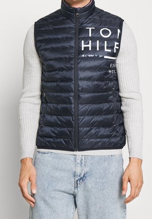 Homme portant un gilet matelassé noir avec le texte « Tom Hilf » sur un pull côtelé gris clair et un jean bleu clair, debout avec les mains relâchées.