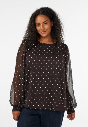 Woman with curly hair wearing black sheer long-sleeve blouse with white polka dots and dark blue jeans, smiling slightly.