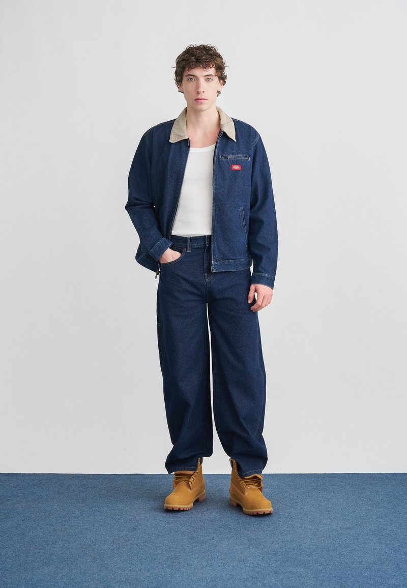 Veste en jean avec col beige, jean bleu et bottes couleur tan. La tenue présente une coupe décontractée et un logo rouge sur la veste.