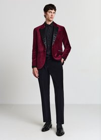 Weinroter Samt-Blazer mit schwarzen Satin-Revers, kombiniert mit einem schwarzen Hemd und maßgeschneiderten schwarzen Hosen. Glänzende schwarze Anzugschuhe.