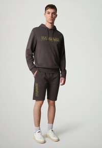 Felpa nera con cappuccio e tasca frontale, con testo grafico giallo, abbinata a pantaloni corti coordinati con branding laterale. Sneakers bianche e calze grigie.