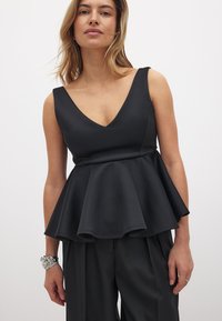 Žena v bezrúchovom čiernom peplum topi s hlbokým výstrihom do V a čiernych širokých nohaviciach, doplnená o strieborný náramok a prsteň.