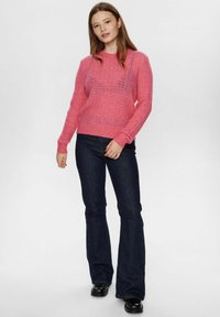 Maglione rosa lavorato a maglia con un design testurizzato, caratterizzato da una lavorazione a rete e polsini a costine, abbinato a jeans svasati scuri e stivali neri.