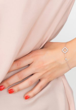 Silver bracelet with a flower-shaped design and clear stones, featuring a thin chain and a small round accent stone. Smooth fabric background.