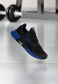 Μαύρο και μπλε αθλητικό παπούτσι Reebok που αιωρείται πάνω από έναν γυαλιστερό δάπεδο γυμναστηρίου με θολούς αλτήρες στο φόντο.