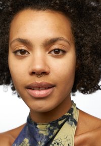 Portrait en gros plan d'une personne avec des cheveux bouclés et un anneau septum en or. Elle porte un haut sans manches marbré vert et bleu. Texture de peau lisse, tons neutres.