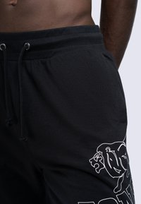 Un survêtement noir présente une taille avec cordon et un design graphique d'un lion sur la jambe gauche. Le matériau semble doux et sans texture.