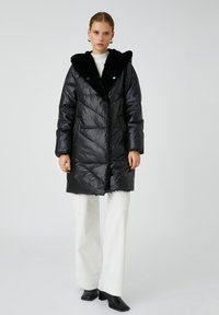 Vrouw in een zwart gewatteerd winterjack met een bontomzoomde capuchon, witte wijde broek, zwarte hoge laarzen en gouden oorringen, staande op een grijze achtergrond.