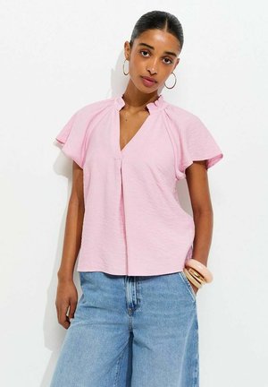 Młoda kobieta ubrana w jasnoróżową bluzkę z krótkim rękawem i falbankowym kołnierzem, niebieskie dżinsy, kolczyki koła i bransoletki na prawym nadgarstku.