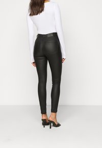 Black faux leather skinny jeans with back pockets, high waist, and ankle length, paired with black heeled slippers and a white long-sleeve shirt.