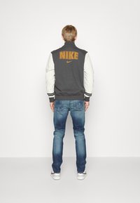 Donkergrijze varsity-jack van Nike met witte mouwen, voorzien van grote oranje "NIKE"-tekst op de achterkant, gecombineerd met blauwe jeans.