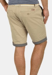 Khaki shorts med opfoldede grå kanter, der har to baglommer, et vævet bælte med gule accenter og et lille logo mærke bagpå.