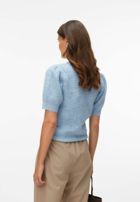 Pull bleu clair en maille avec manches courtes bouffantes et motif texturé, associé à un pantalon beige et un petit sac noir.