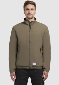 Olivengrøn, zip-up jakke med høj krave, to sidelommer med lynlås og en struktureret stof. Har et logo patch nær kanten.