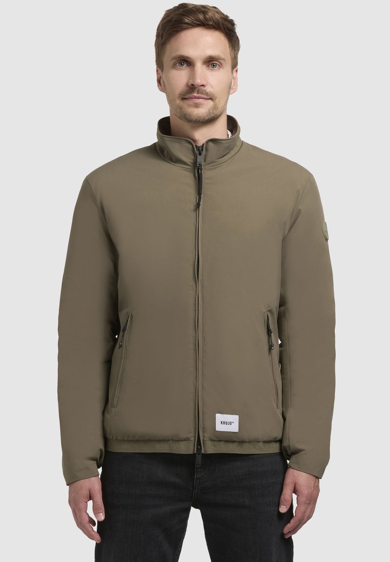 Olivengrøn, zip-up jakke med høj krave, to sidelommer med lynlås og en struktureret stof. Har et logo patch nær kanten.