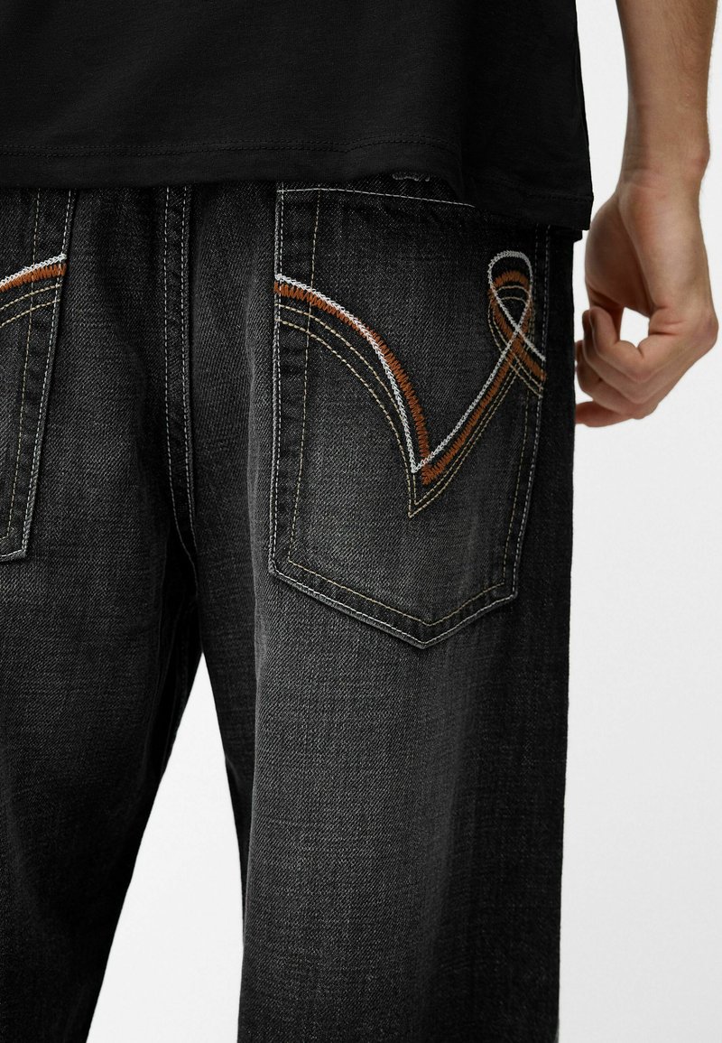 Jeans en denim foncé avec une finition texturée, des coutures orange contrastantes sur des poches arrière en forme, avec une coupe décontractée.