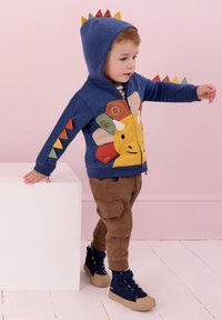Blå zip-up luvtröja med dinosauriedesign, färgglada taggar och applikédetaljer, kombinerad med bruna cargo-byxor och marinblå högstövelsnickers.