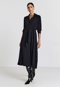 Schwarzes langärmliges Kleid mit Kragen, tailliertem Schnitt und schwingendem Rock. Getragen mit hohen schwarzen glänzenden Stiefeln und einer geschichteten Halskette.