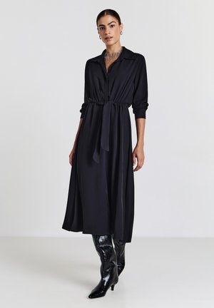 Schwarzes langärmliges Kleid mit Kragen, tailliertem Schnitt und schwingendem Rock. Getragen mit hohen schwarzen glänzenden Stiefeln und einer geschichteten Halskette.