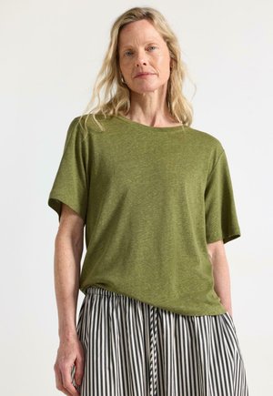 Donna con lunghi capelli biondi che indossa una maglietta verde oliva a maniche corte e una gonna a righe verticali bianca e nera, in piedi davanti a uno sfondo semplice.