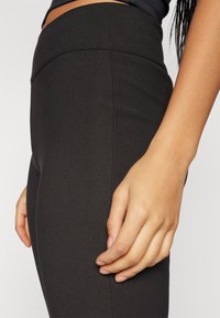 Leggings taille haute noires avec une texture lisse et des détails de couture sur le côté, montrés avec les mains placées le long de la hanche.