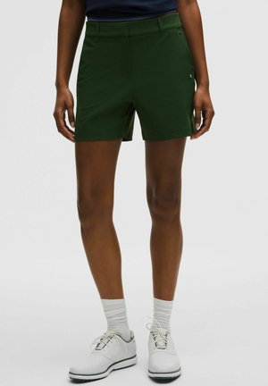 Grüne Shorts aus Stretchstoff mit tailliertem Schnitt, Fronttaschen und einer glatten Textur. Kombiniert mit weißen Schuhen und Socken.