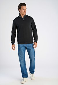 Pullover nero con zip in un tessuto morbido, abbinato a jeans in denim azzurro chiaro e sneakers bianche, creando un outfit casual.