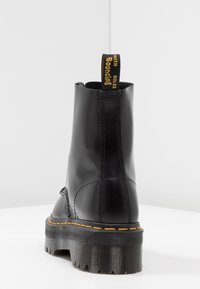 Botín negro de tobillo hecho de cuero brillante con costuras amarillas y una pestaña de agarre que presenta texto negro. Suela de goma gruesa y estriada.