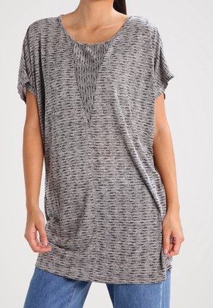 Femme portant un haut ample gris à manches courtes avec des traits noirs et un motif en forme de V près de l'encolure, associé à un jean bleu.