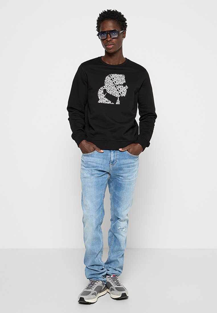 Felpa nera con una grafica sulla parte anteriore, jeans blu chiaro e scarpe da ginnastica grigie. Il modello indossa occhiali da sole scuri.